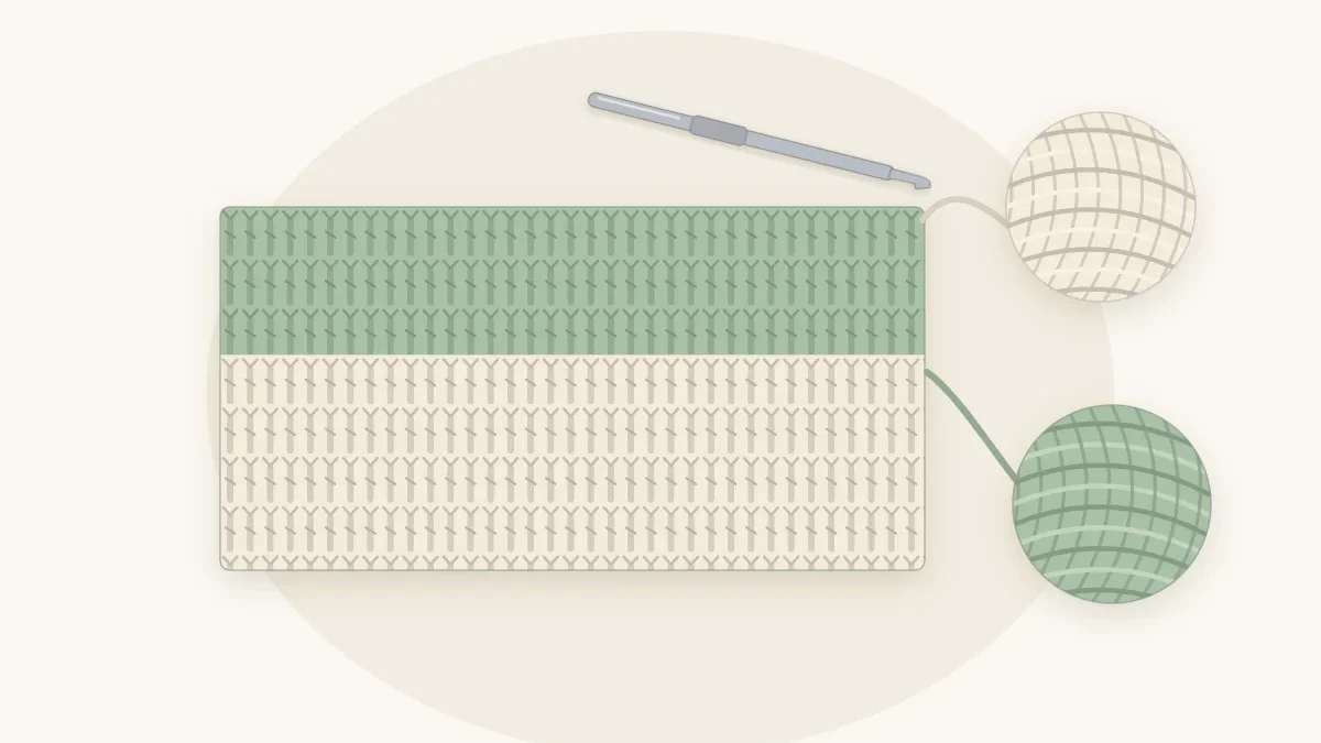 A crochet blanket in progress where a sage green row meets a cream row, with both yarn balls attached and a hook in the work