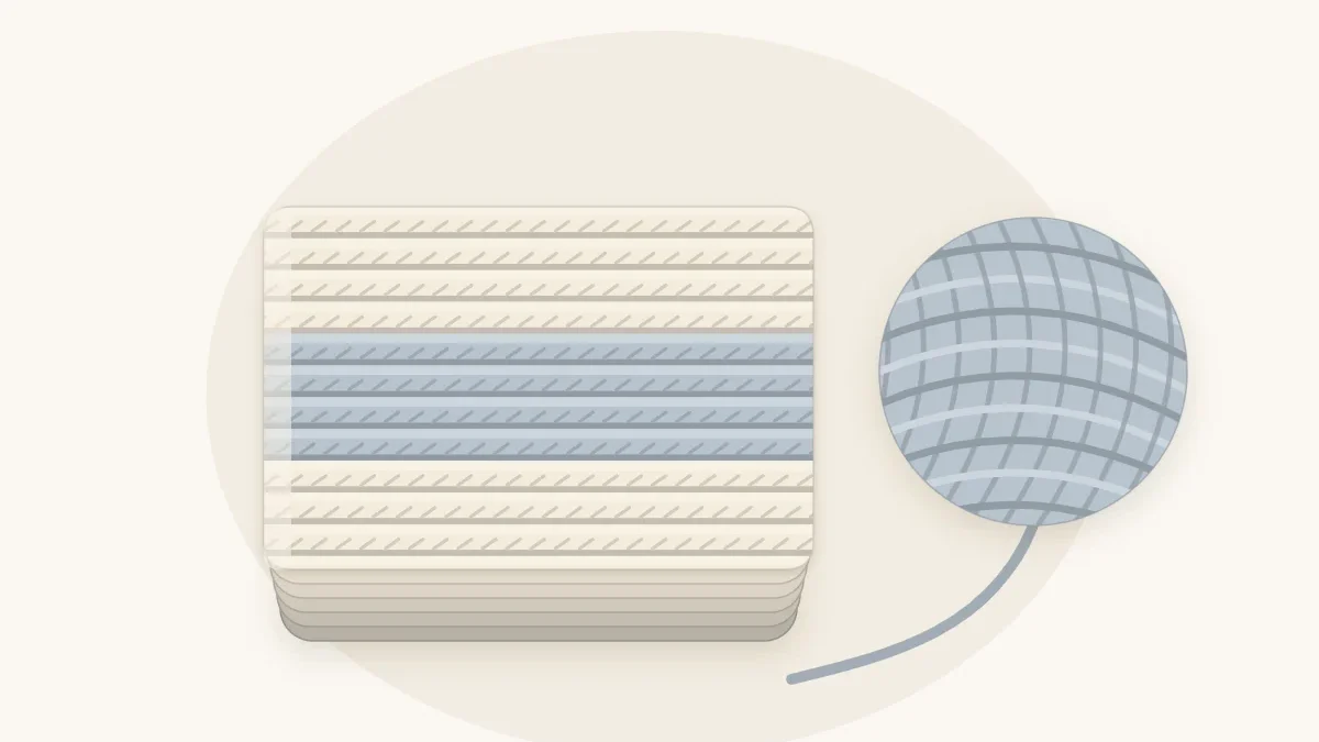 A thick ribbed crochet baby blanket in cream and soft gray-blue folded into a chunky stack beside a large ball of bulky yarn